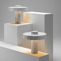 Two sculptural white marble pillar table lamps displayed on tiered white pedestals with warm ambient light glowing from the base