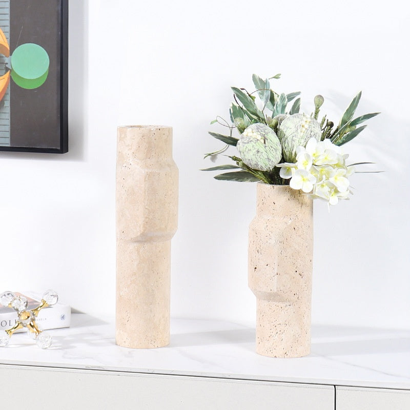 Two different sized travertine stone sculptural vases styled on a white marble surface with green foliage and white flowers