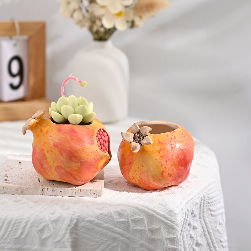 Two sculptural pomegranate ceramic planters in ochre and red tones styled on a white lace tablecloth