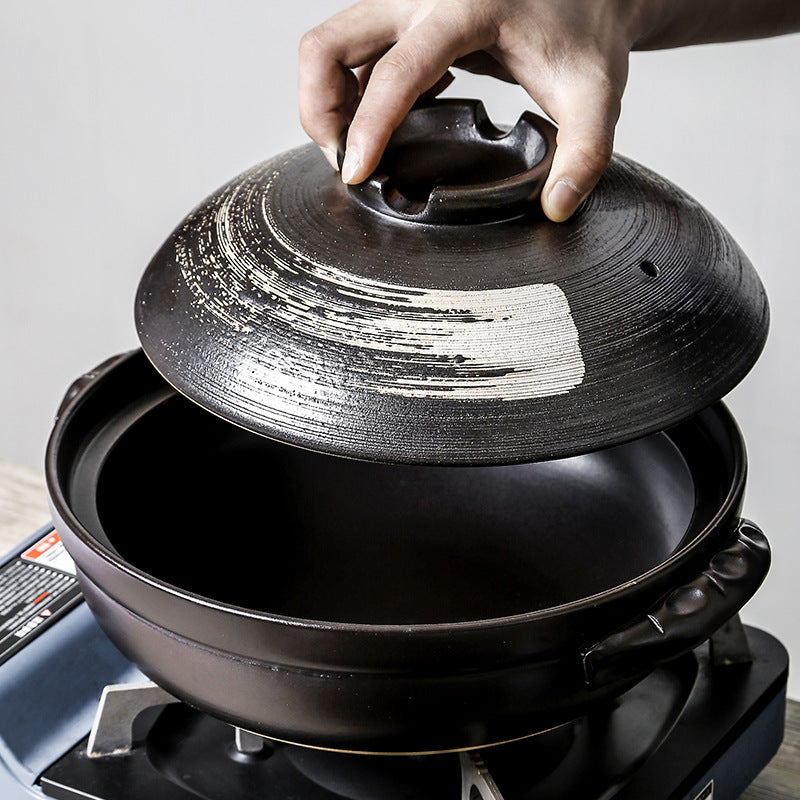 Hand lifting the vented lid of a matte black ceramic pot resting on a portable gas burner