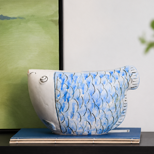Blue and white ceramic fish vase displayed on books with green artwork in the background