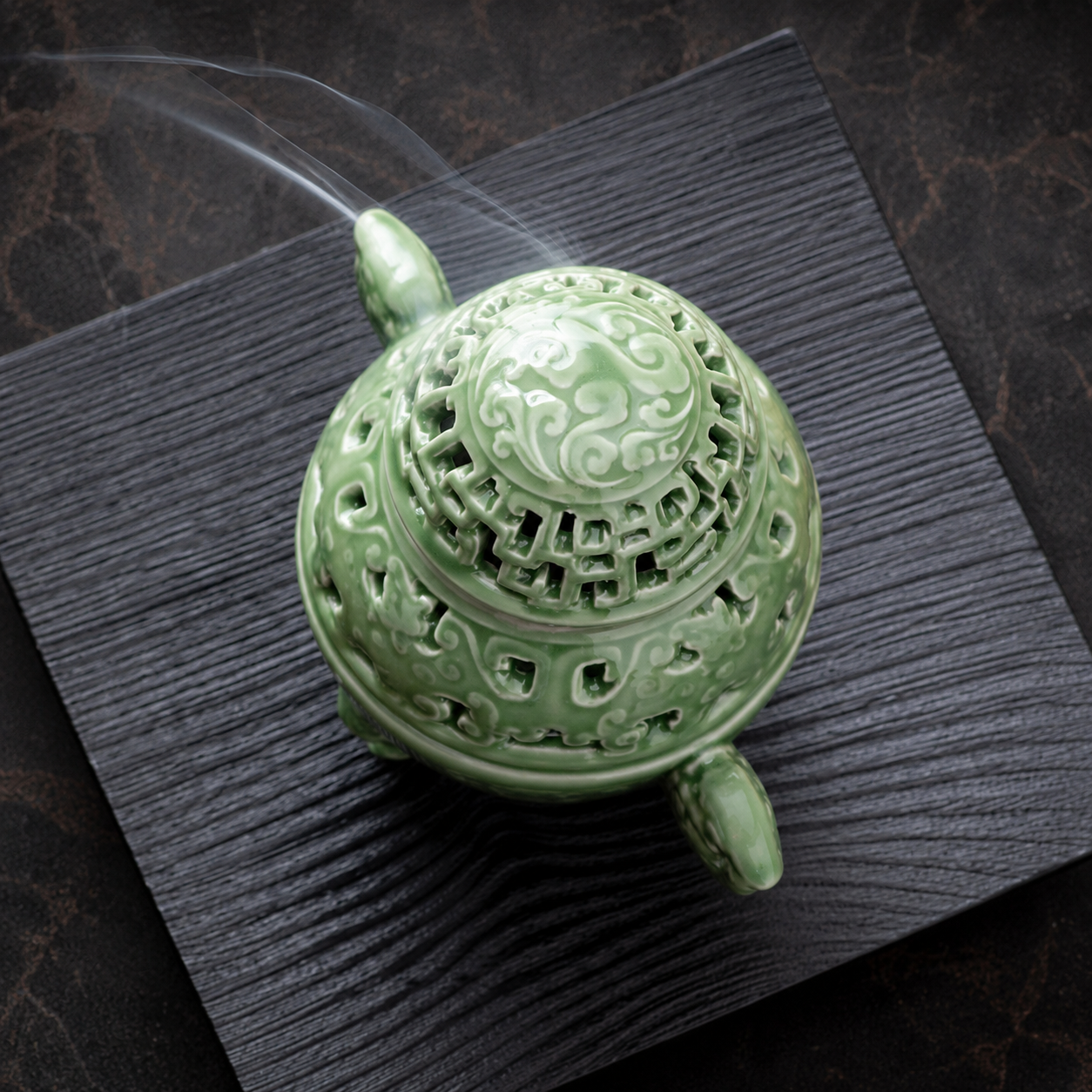 Top view of a celadon green ceramic incense burner releasing smoke on a dark tray