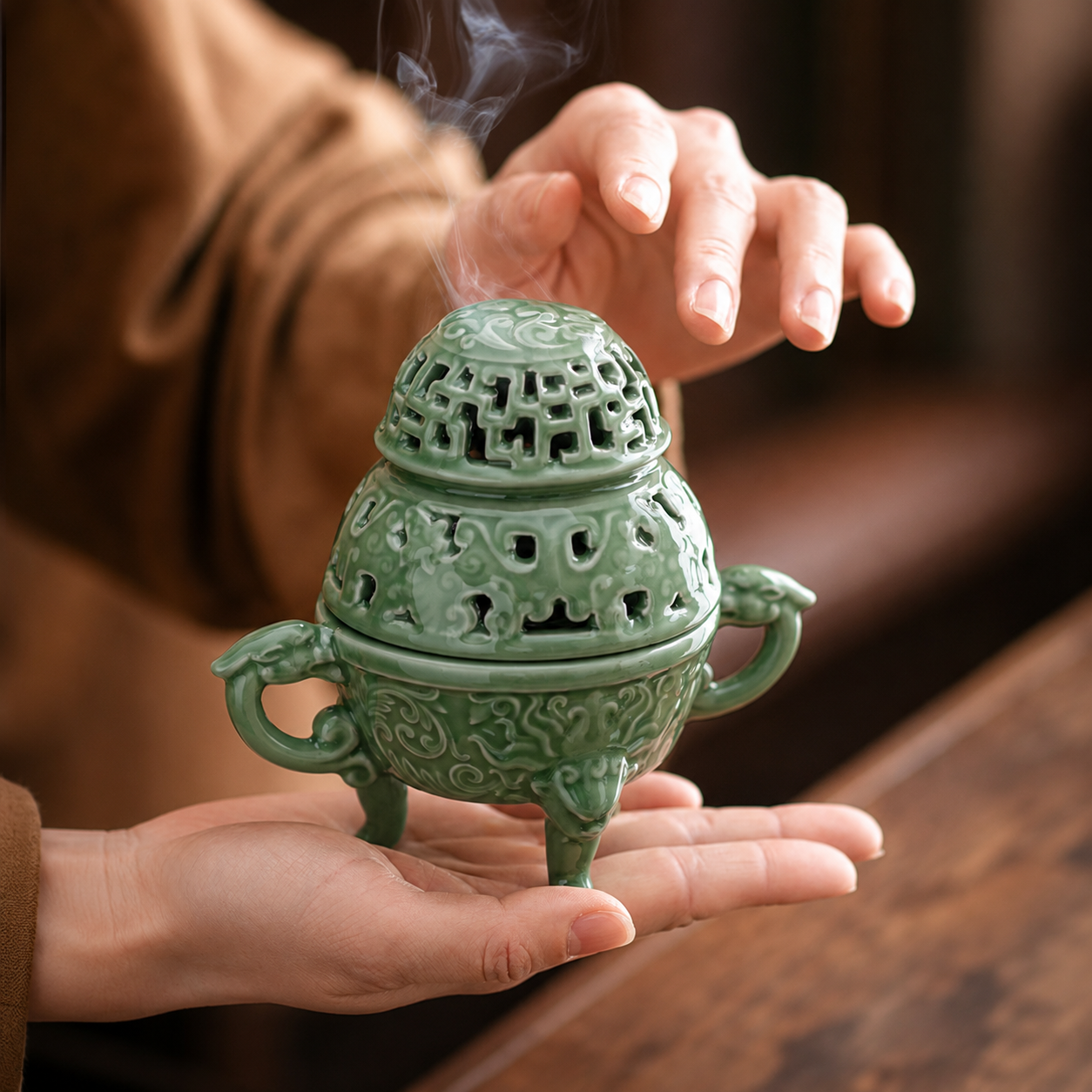 Hands holding a celadon green ceramic incense burner with smoke rising from the lid