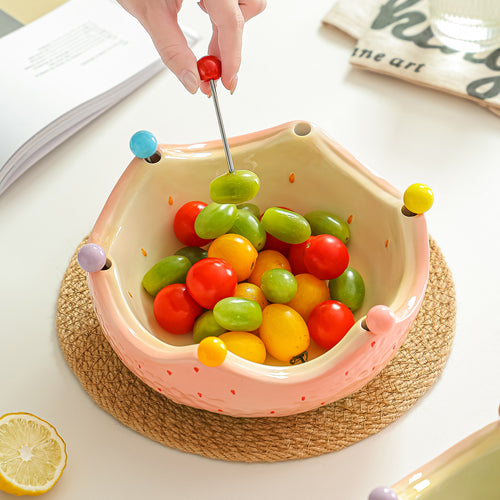 Pink ceramic crown fruit bowl filled with colorful fruit on a woven placemat with a fruit fork lifting a grape