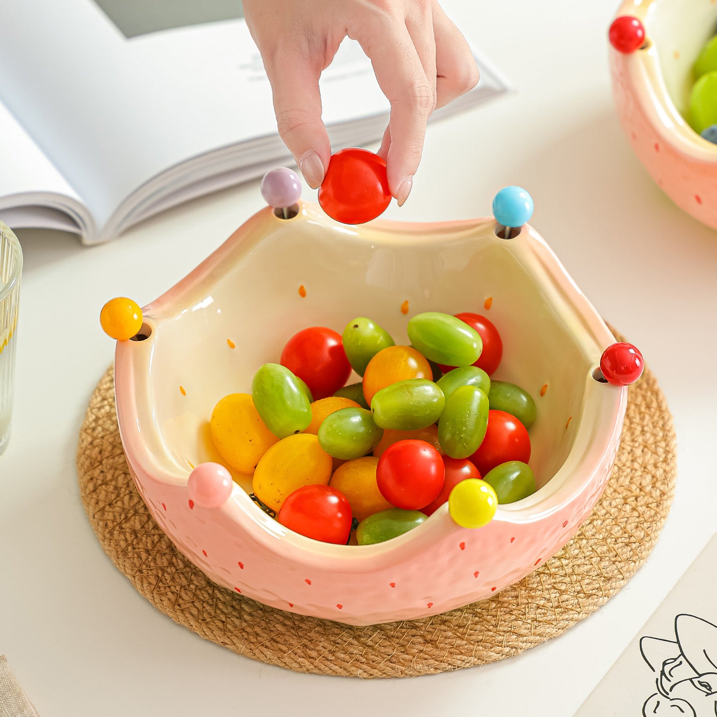 Pink ceramic crown fruit bowl filled with colorful fruit on a woven placemat with a hand using a fruit fork