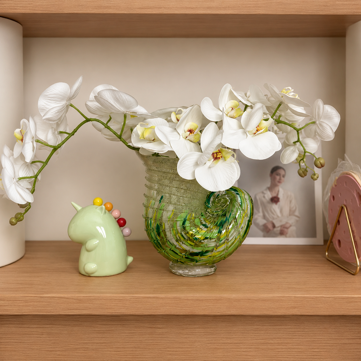 Green and amber nautilus shell art glass vase with white orchids on wooden shelf