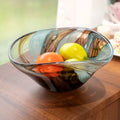 Hand-blown swirl art glass fruit bowl with blue and amber-brown tones styled with fresh fruit on a wooden table