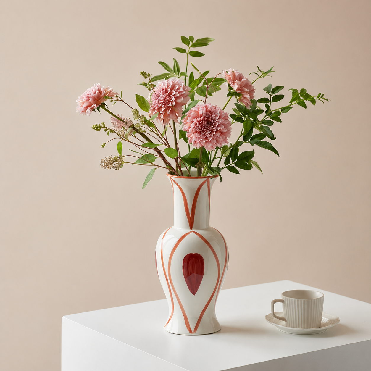 Hand painted heart ceramic flower vase with pink dahlias and greenery on white table beside cup