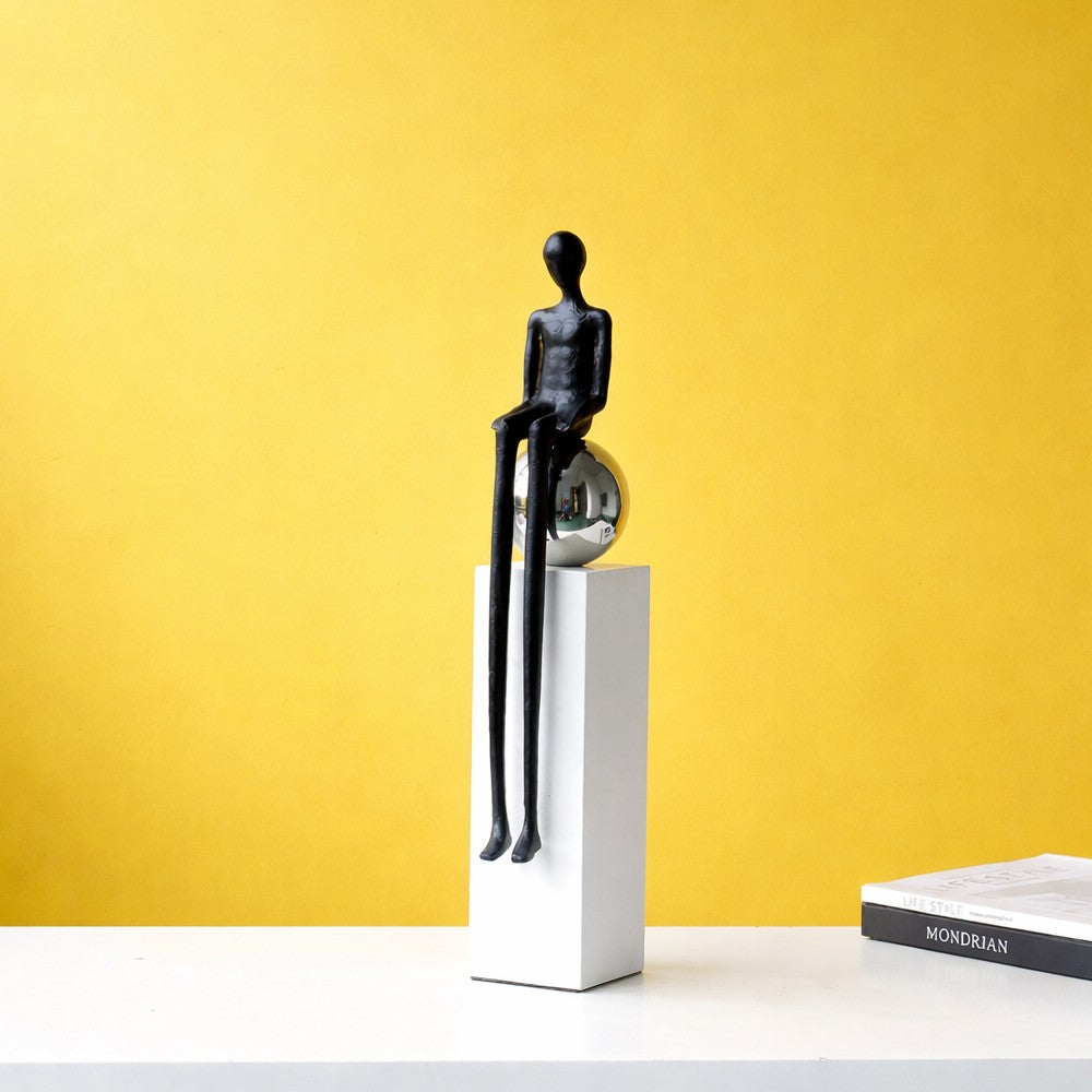 Black sitting abstract figure sculpture on white pedestal with reflective chrome sphere detail