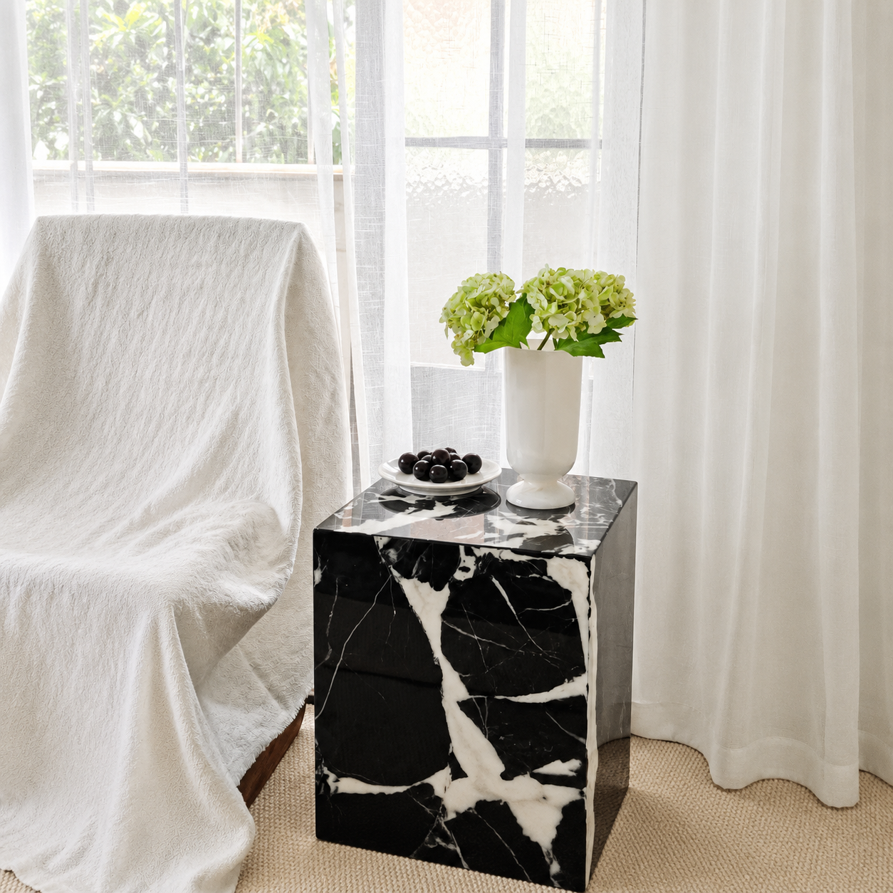 White marble pedestal flower vase with green hydrangeas on black marble side table by sheer curtains