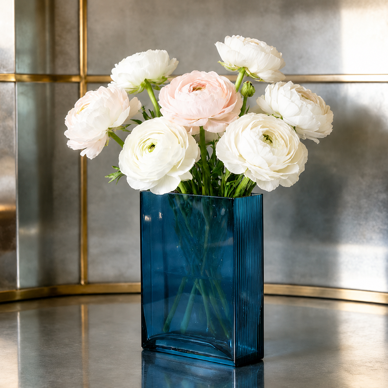Blue rectangular hand blown glass vase with white ranunculus flowers on a metallic table
