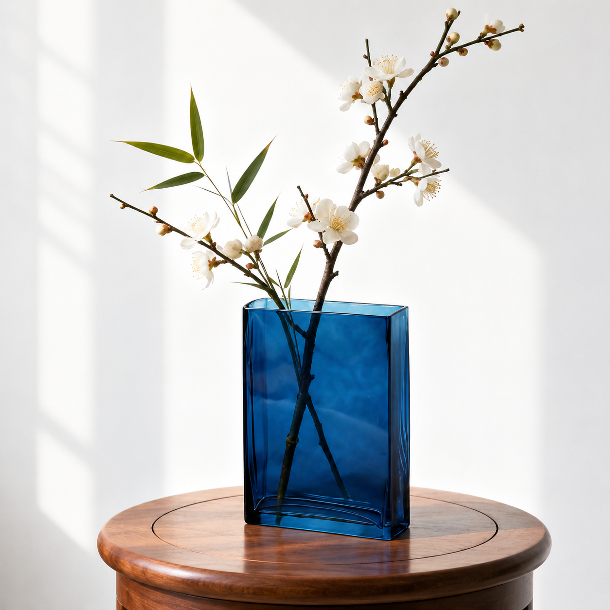 Blue rectangular hand blown glass vase with white blossoms on a round wooden table
