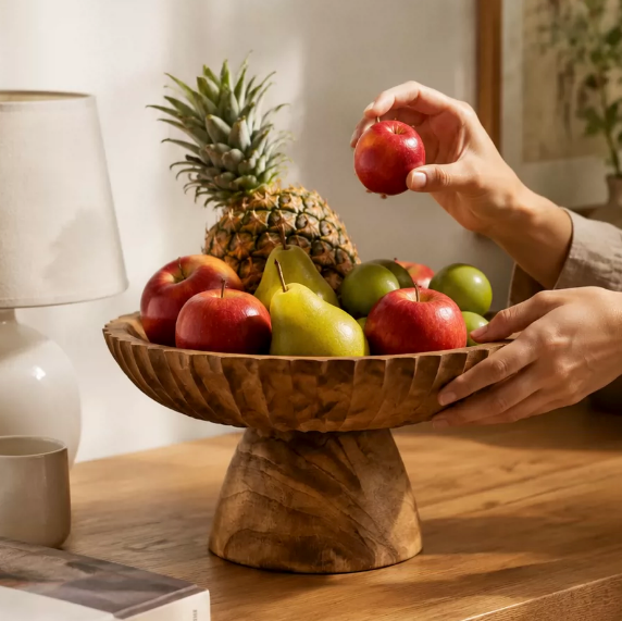 Rustic wood pedestal bowl with assorted fruit and a hand placing a red apple
