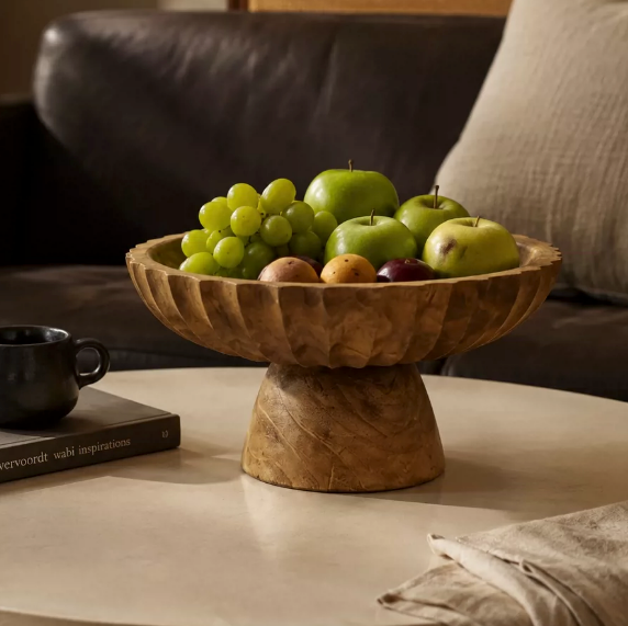 Wooden pedestal fruit bowl filled with green grapes on a light table beside a glass