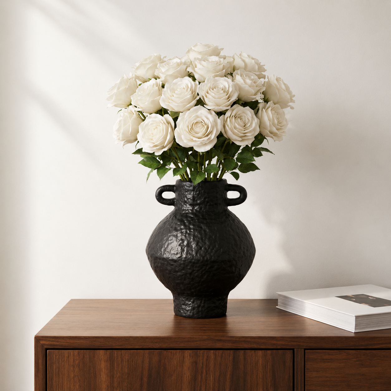 Black textured ceramic vase with white roses displayed on a wooden cabinet