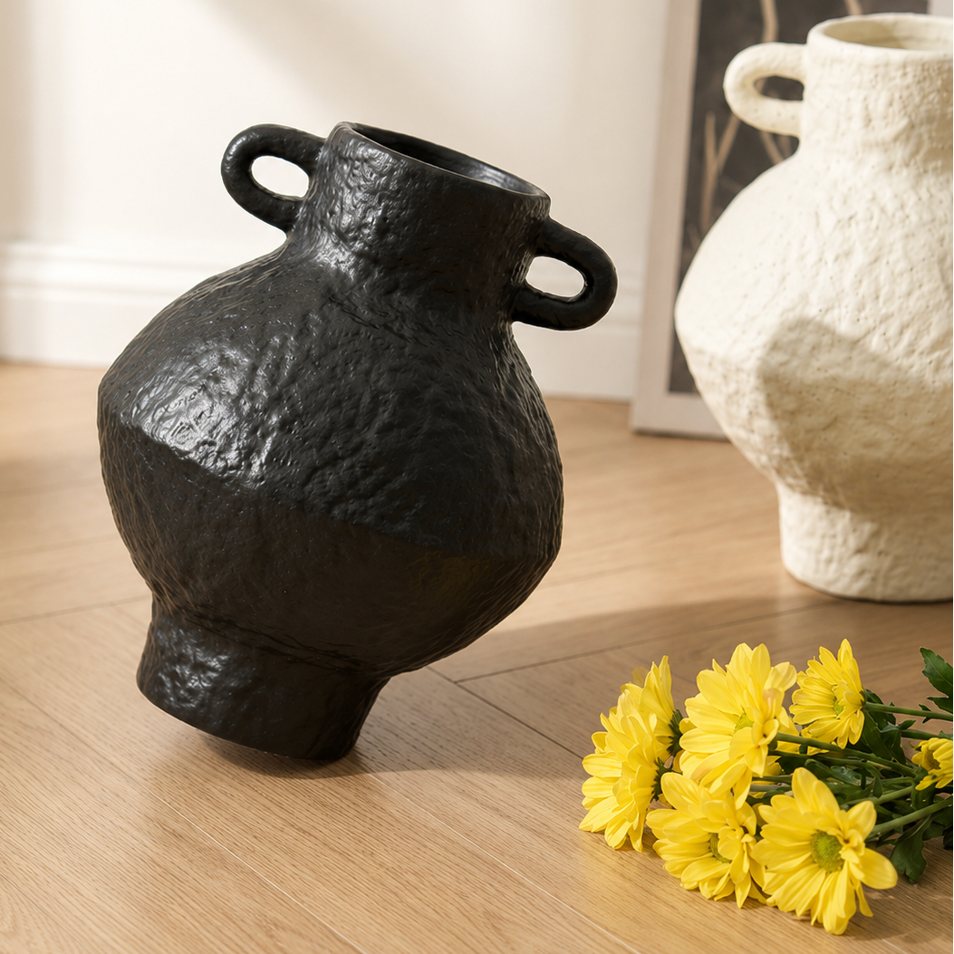 Tilted black ceramic vase with handles on the floor beside yellow flowers and an ivory vase in the background