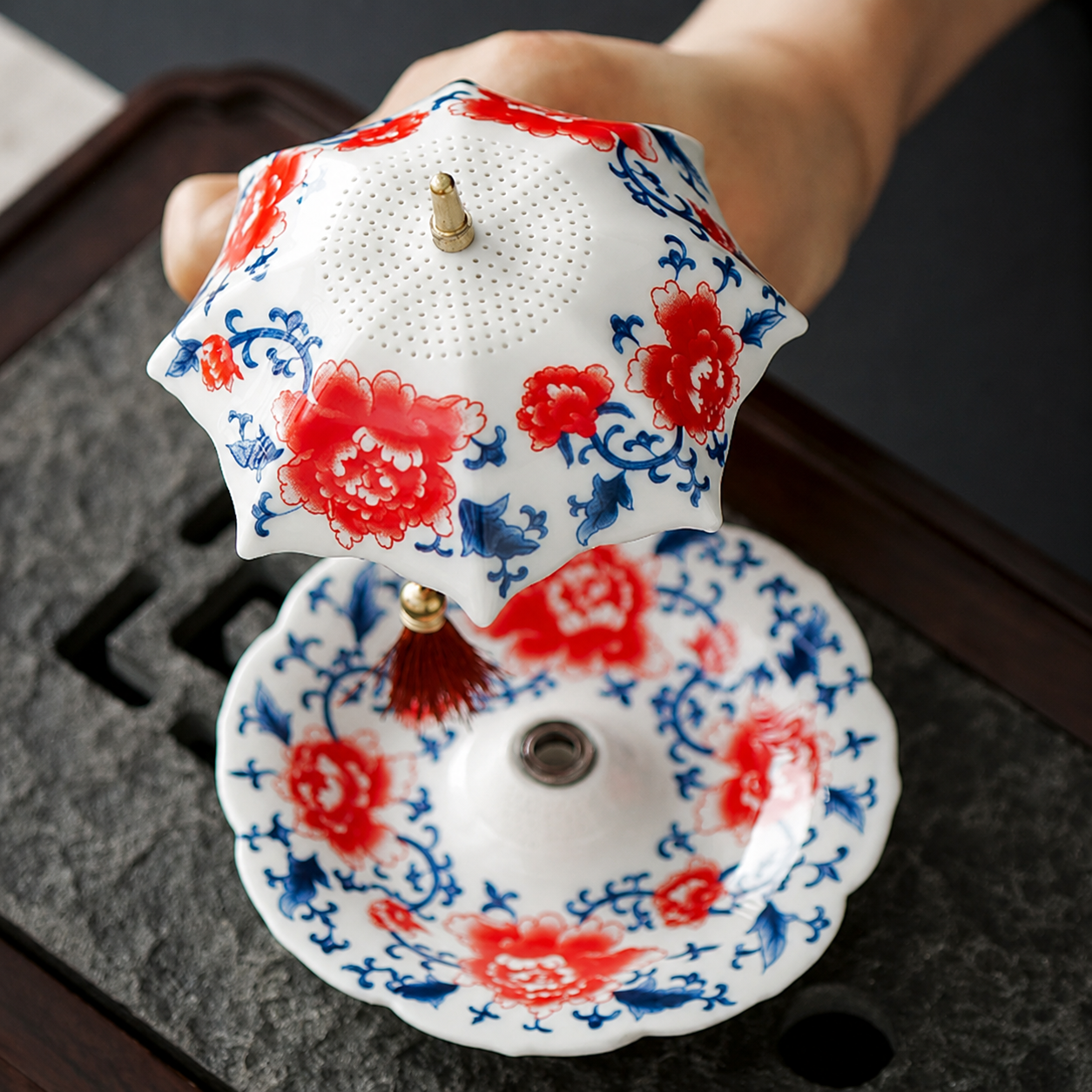 Hand holding red floral ceramic tea strainer above matching drip tray on a tea tray
