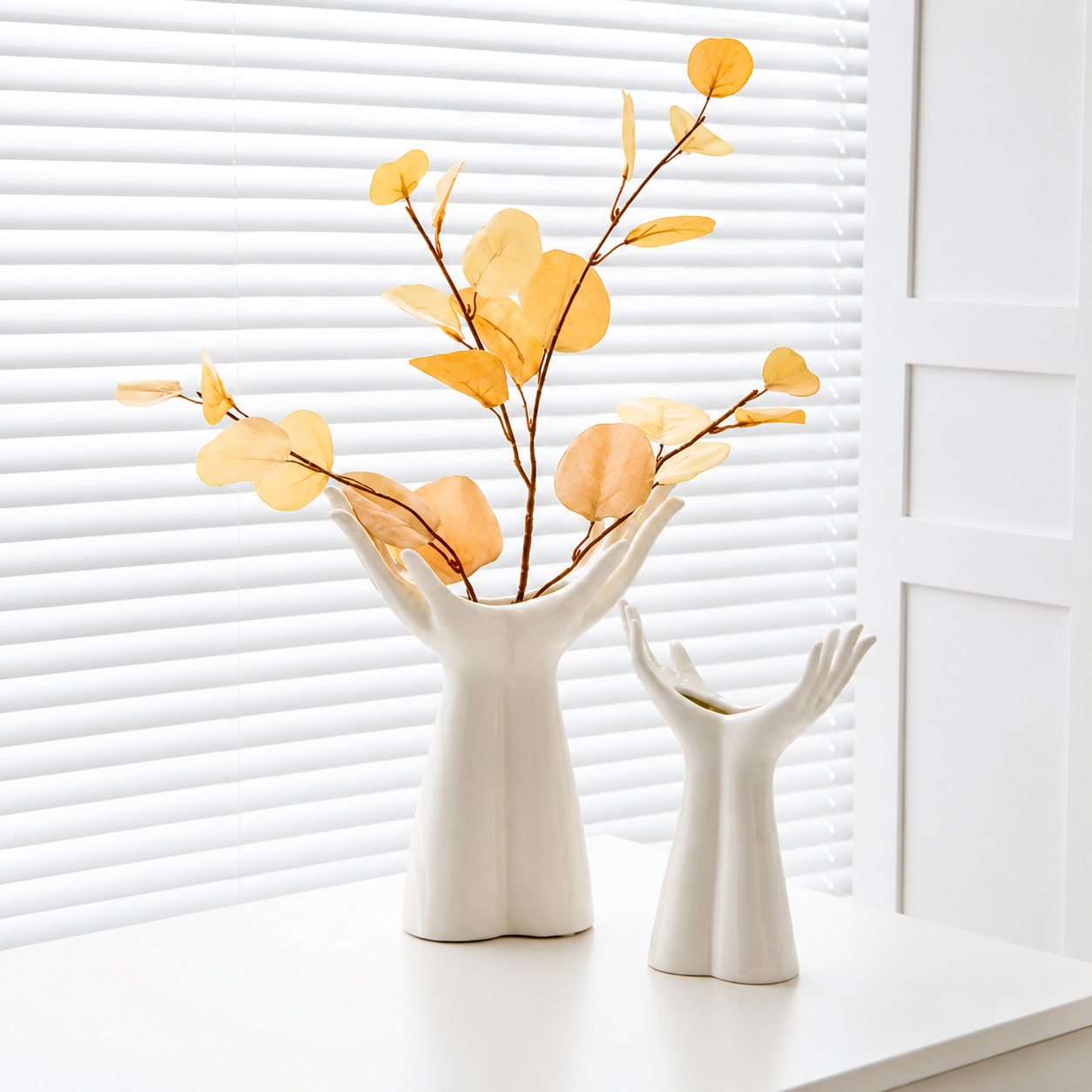 White ceramic open palm vases in two sizes styled with golden botanical stems by a window
