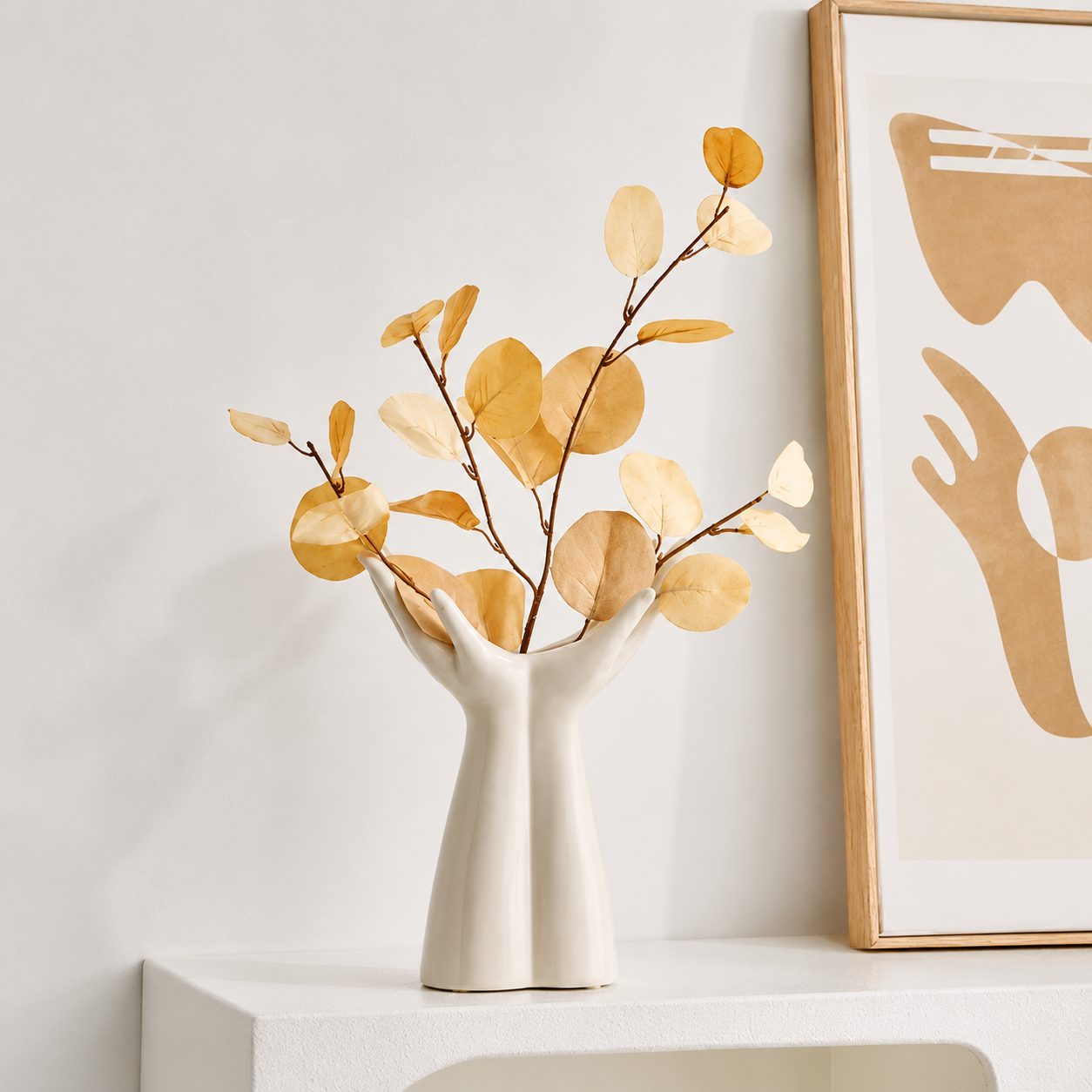 Sculptural white ceramic hand vase with golden leaf stems beside wall art