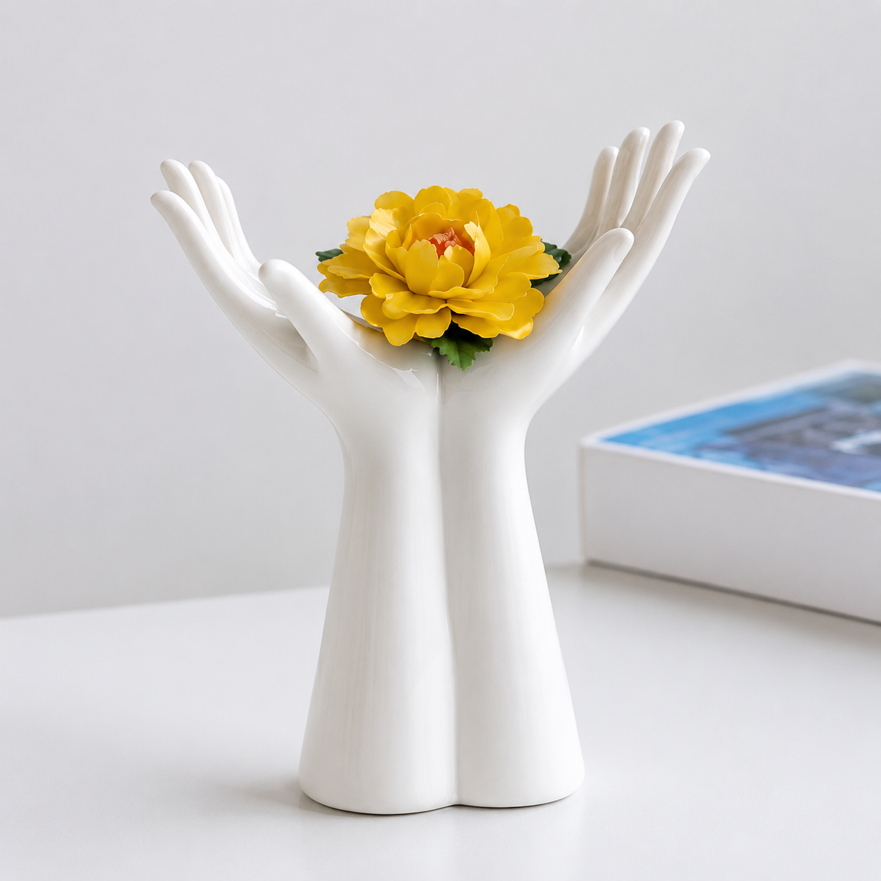 White ceramic open palm vase holding a yellow flower on a minimalist shelf