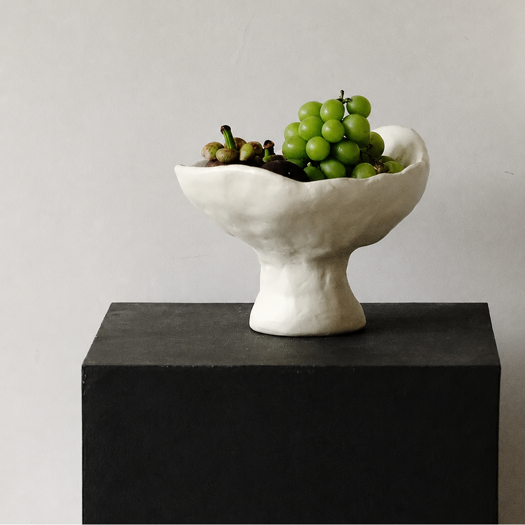 White sculptural ceramic pedestal bowl with green grapes on black display plinth