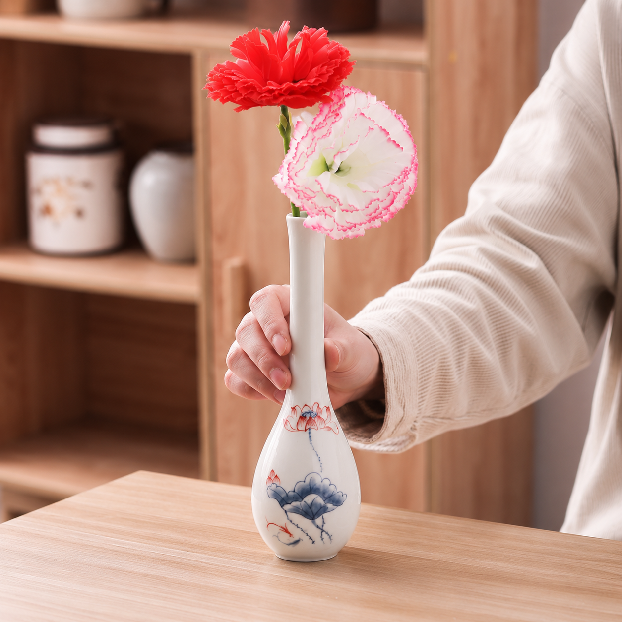 Hand holding a slim ceramic bud vase with blue lotus artwork and two flowers on a wooden table