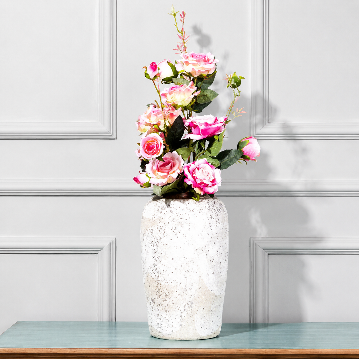 Tall speckled white ceramic vase with bronze rim holding pink roses against a paneled wall