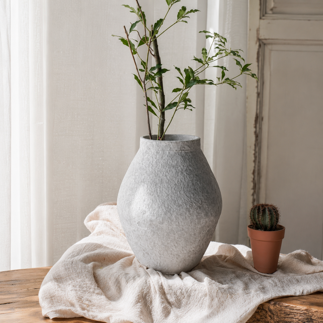 Tall textured ceramic vase with leafy stems beside small cactus on soft linen cloth