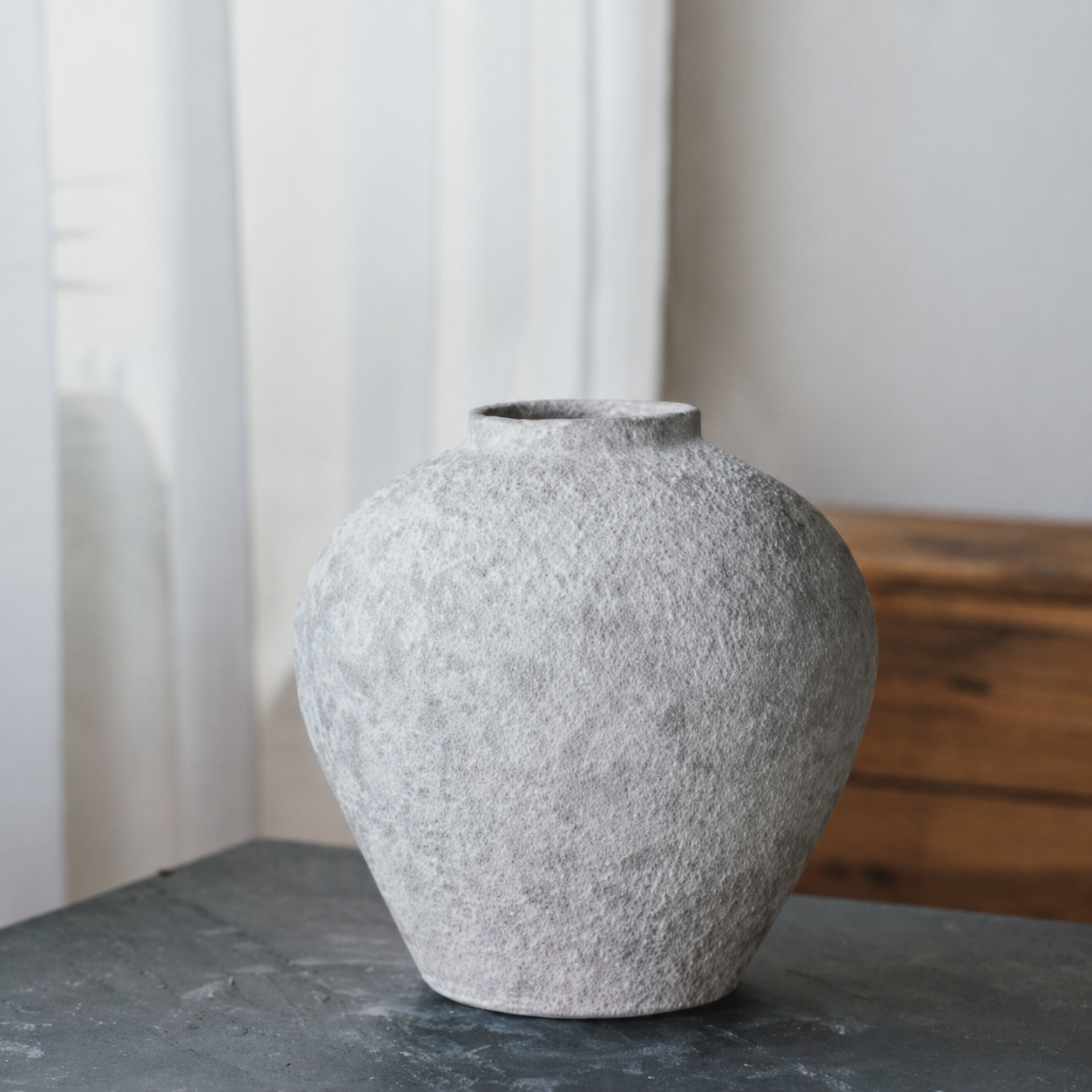 Round textured ceramic vase with stone finish on dark tabletop near window