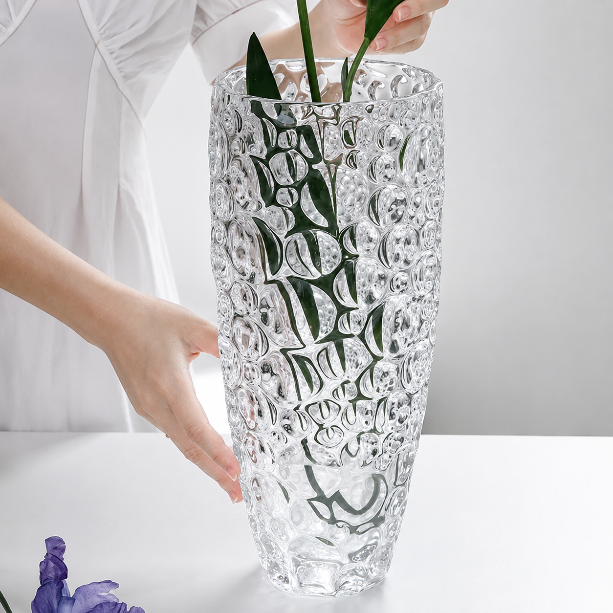 Person arranging green stems inside a tall clear bubble glass vase