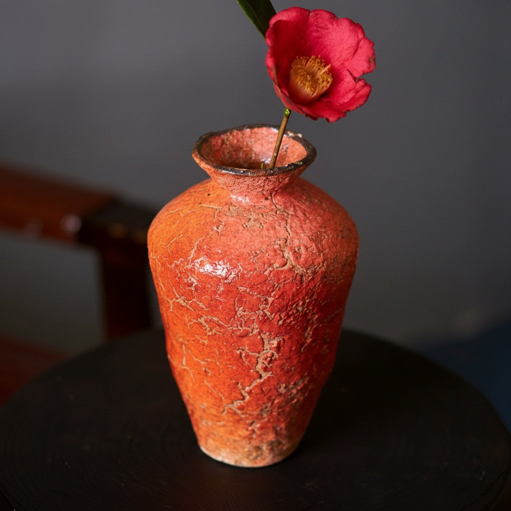 Tall orange ceramic vase with single red flower on dark table
