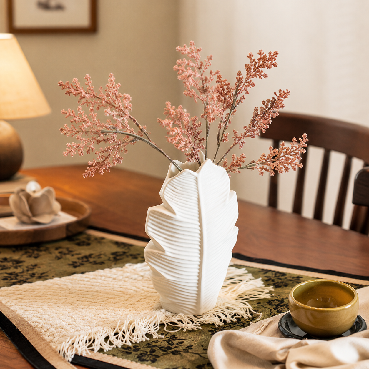 White ceramic leaf vase with pink dried stems in a cozy table setting