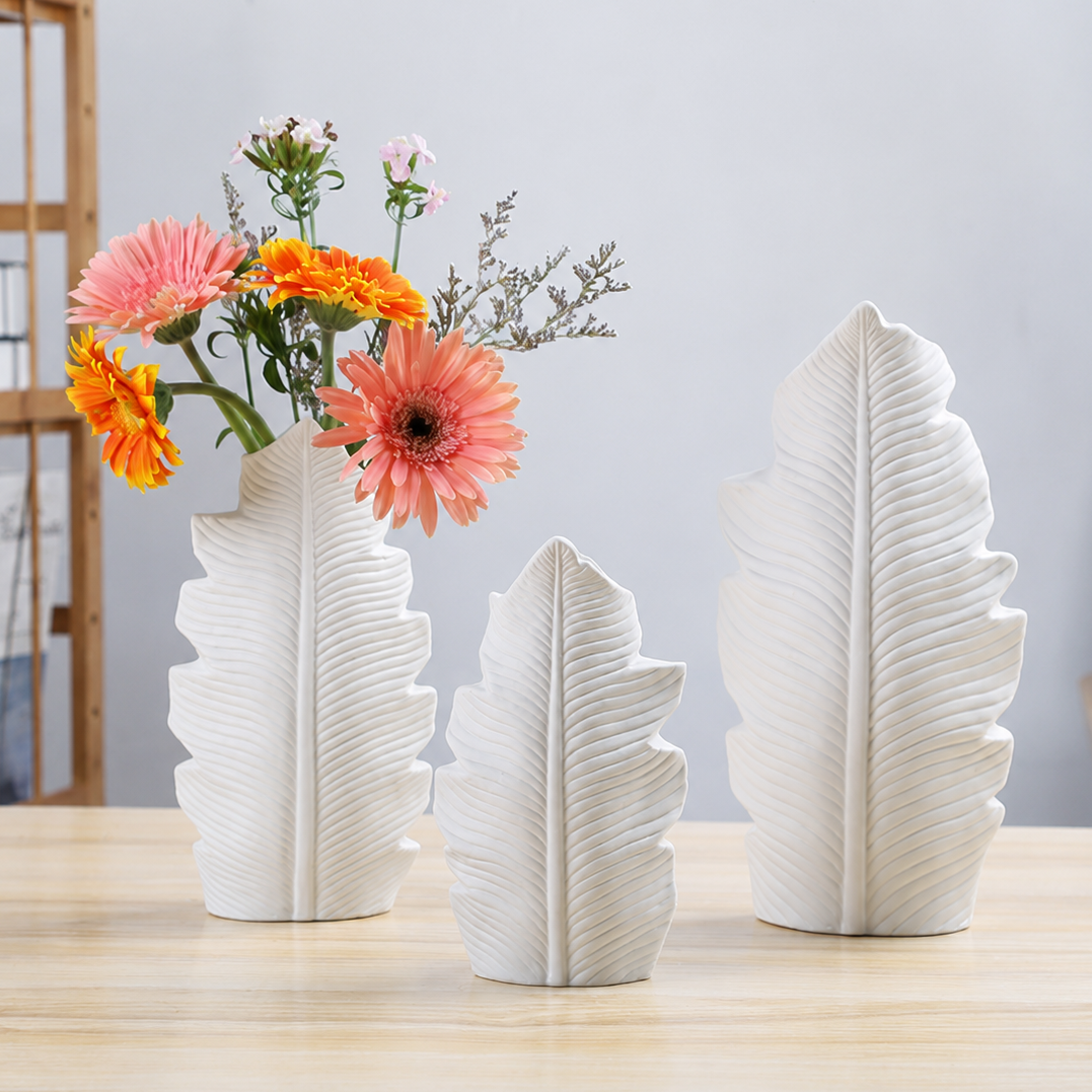 Set of three white ceramic leaf vases with flowers on a light wood table