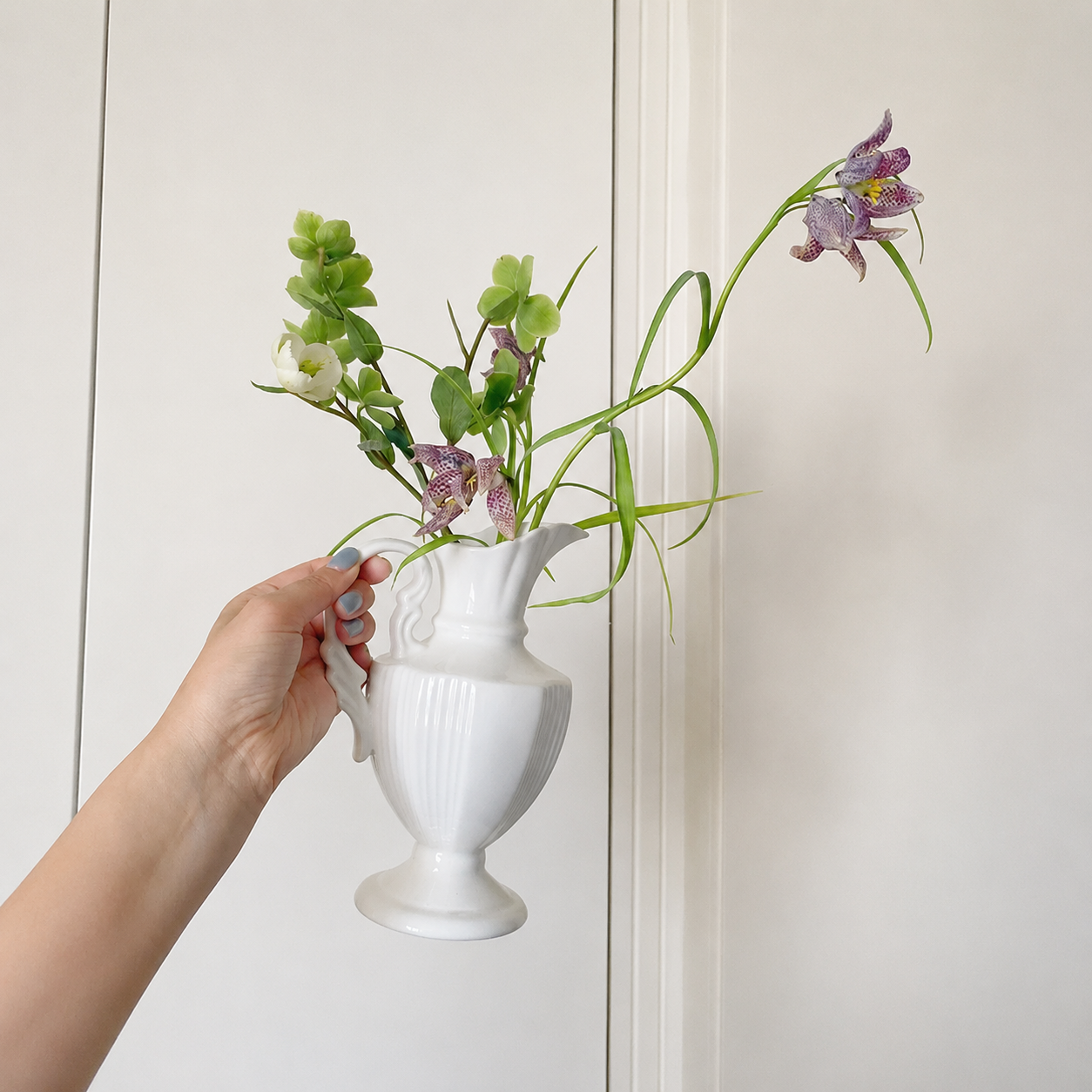 Hand holding white ceramic pitcher vase with purple flowers and green stems