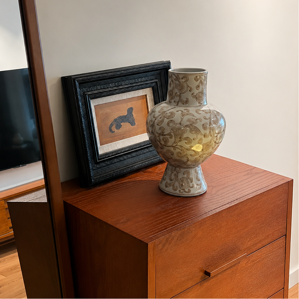 Decorative white and gold ceramic vase displayed on a wooden dresser beside framed artwork and mirror