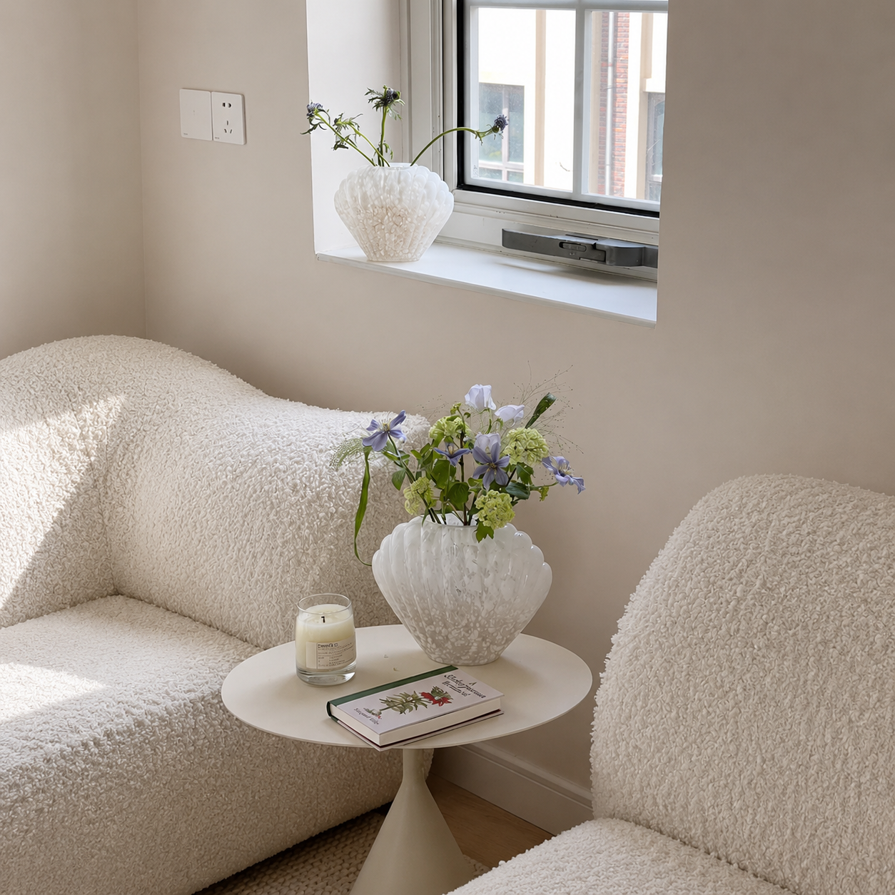White shell-shaped glass vase with flowers on side table between boucle chairs in soft neutral room