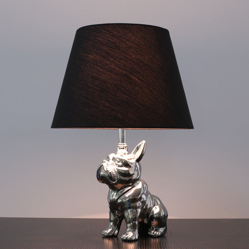 Front view of silver ceramic French bulldog table lamp with black shade lit