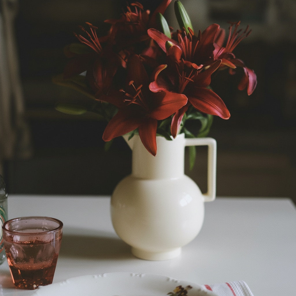 Glossy cream ceramic pitcher holding red lilies on a dining table with glassware