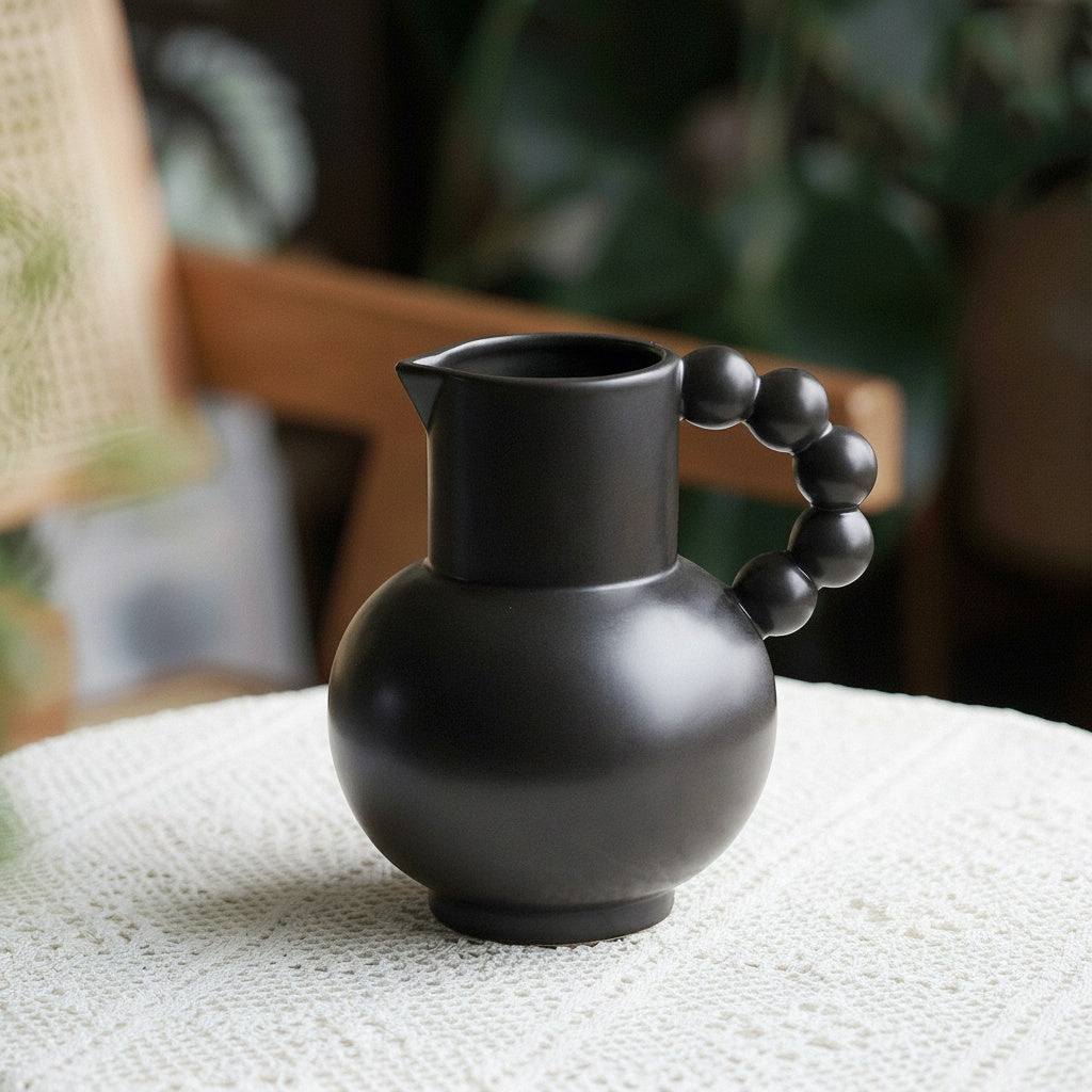 Matte black ceramic pitcher with beaded handle on a lace tablecloth
