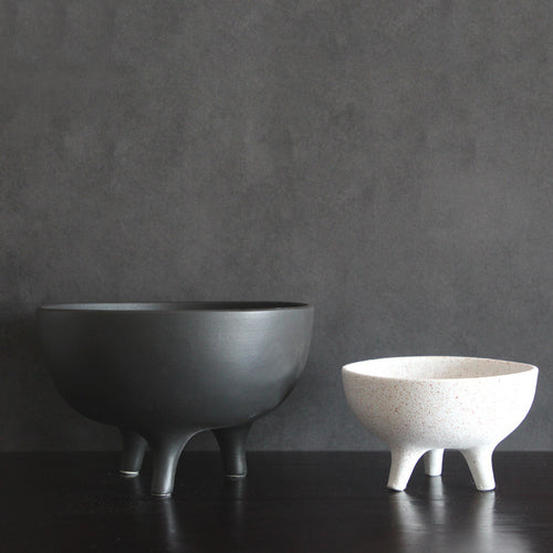 Two modern minimalist ceramic pedestal bowls in contrasting black and white matte finishes against a dark textured background