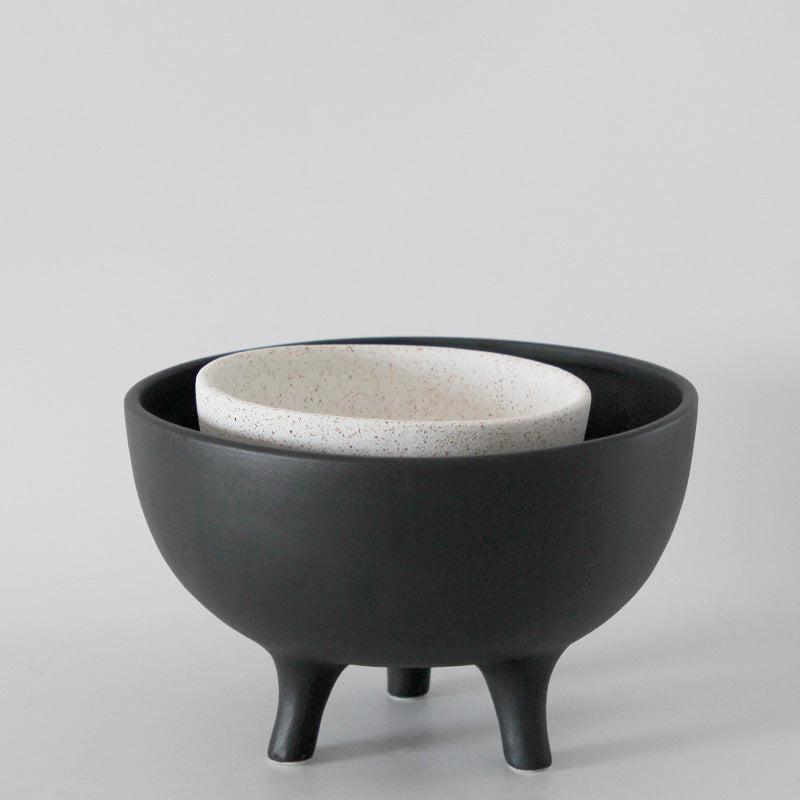 Nesting ceramic tripod bowls showing the speckled cream vessel stacked inside the larger charcoal black pedestal bowl.