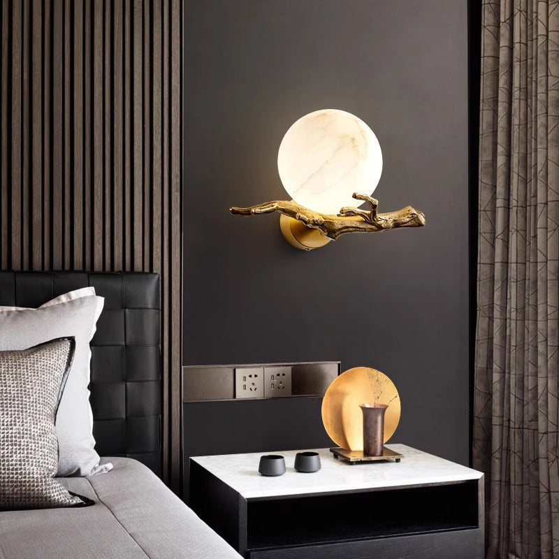 Illuminated alabaster globe wall sconce resting on textured gold branch arm in a dark luxury bedroom