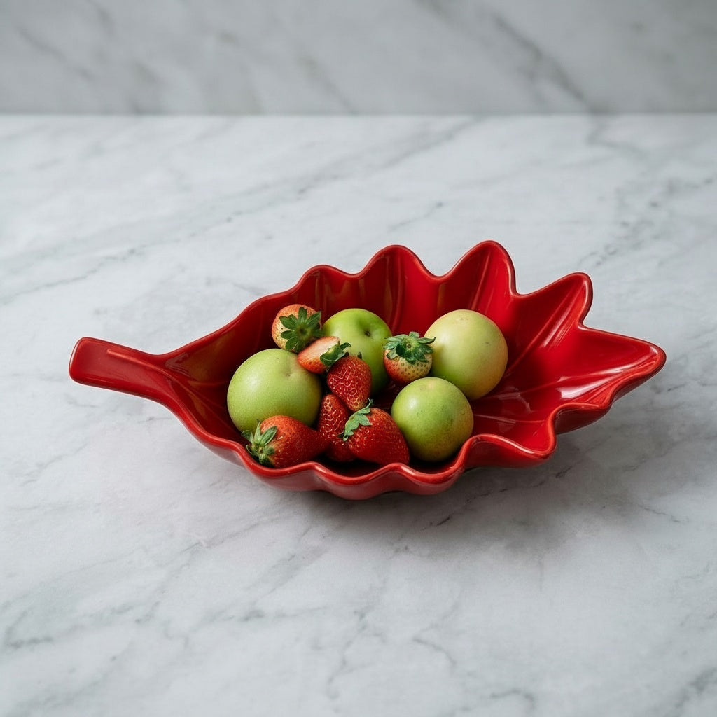 Red glazed ceramic leaf shaped bowl filled with apples and strawberries on a white marble surface