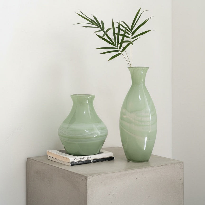 Two sage green marbled glass vases with greenery on a wooden console