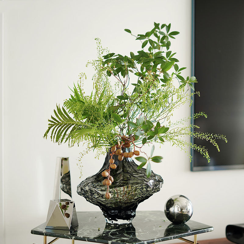 Smoke grey geometric glass vase with green foliage on marble table
