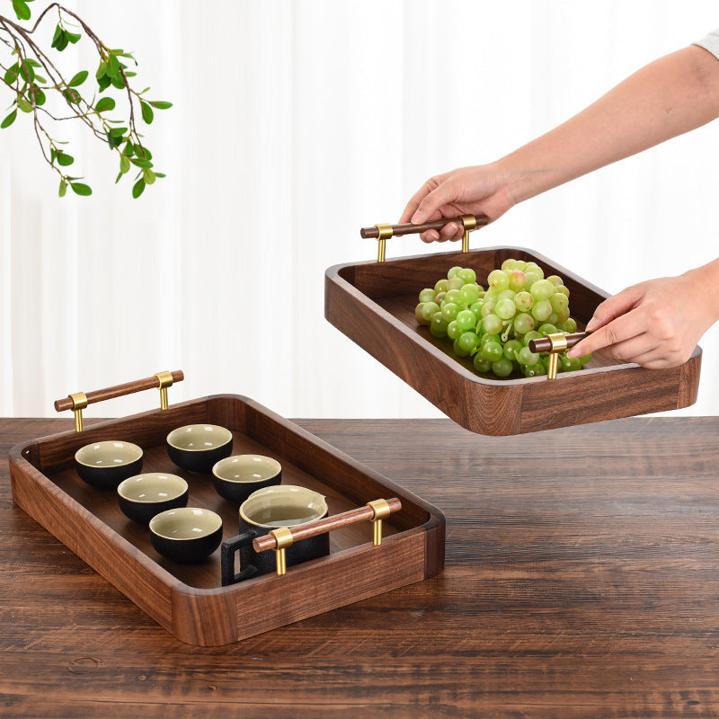 Handcrafted walnut wood serving tray held by gold handles displaying grapes and a tea set in a bright setting