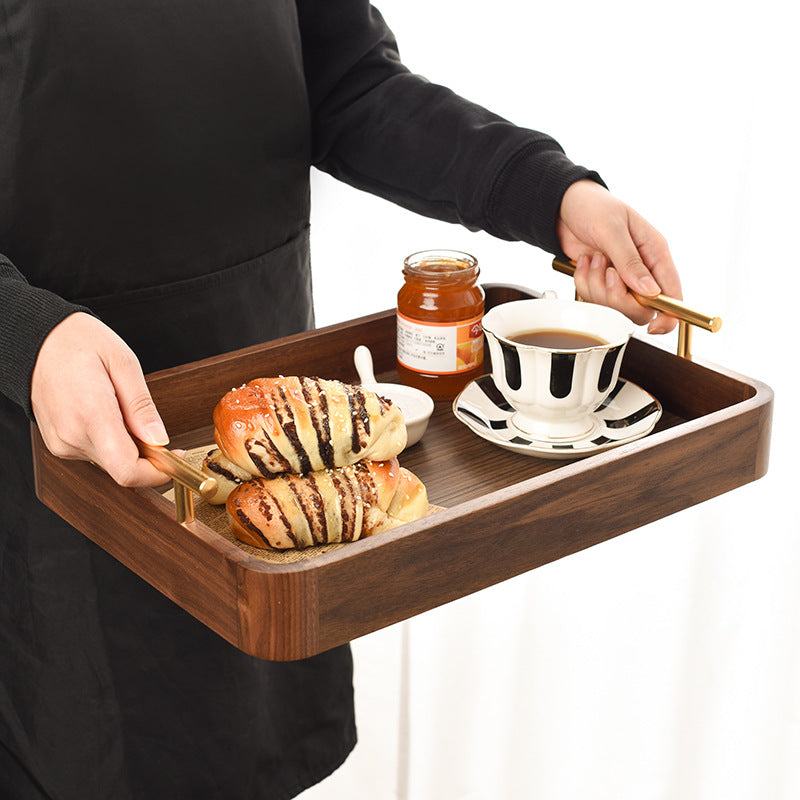 Person holding rectangular wooden serving tray with pastries and coffee cup showing ergonomic brass handles