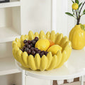 Yellow ceramic banana fruit bowl filled with grapes and lemons on white side table